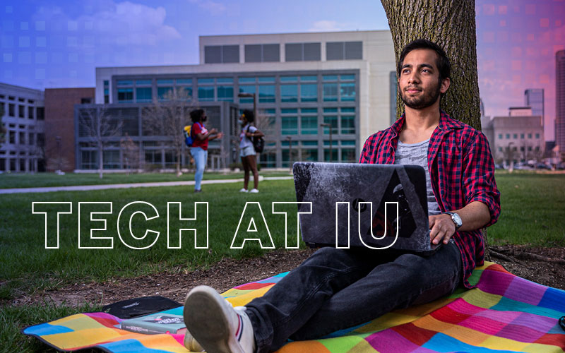 A man seated on the grass with an open laptop. The words tech at IU are superimposed on the image.