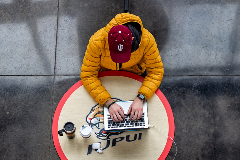 Person sits at IUPUI-branded table, working on a laptop.
